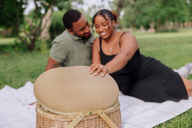 Beige wicker picnic basket with light wood oval lid on white picnic blanket in a sunny park setting.