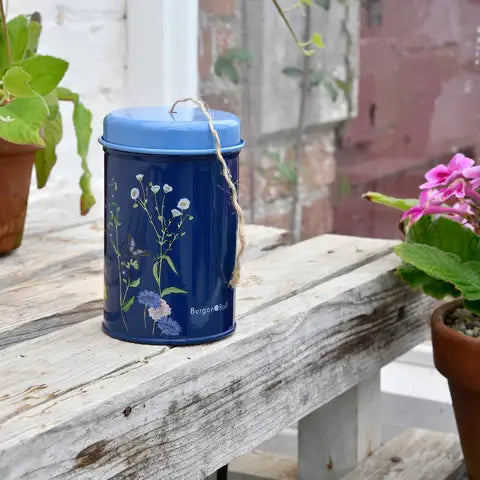 Dark blue cylindrical tin with painted wildflower design and carrying handle, containing jute twine, displayed on weathered wood with potted plants