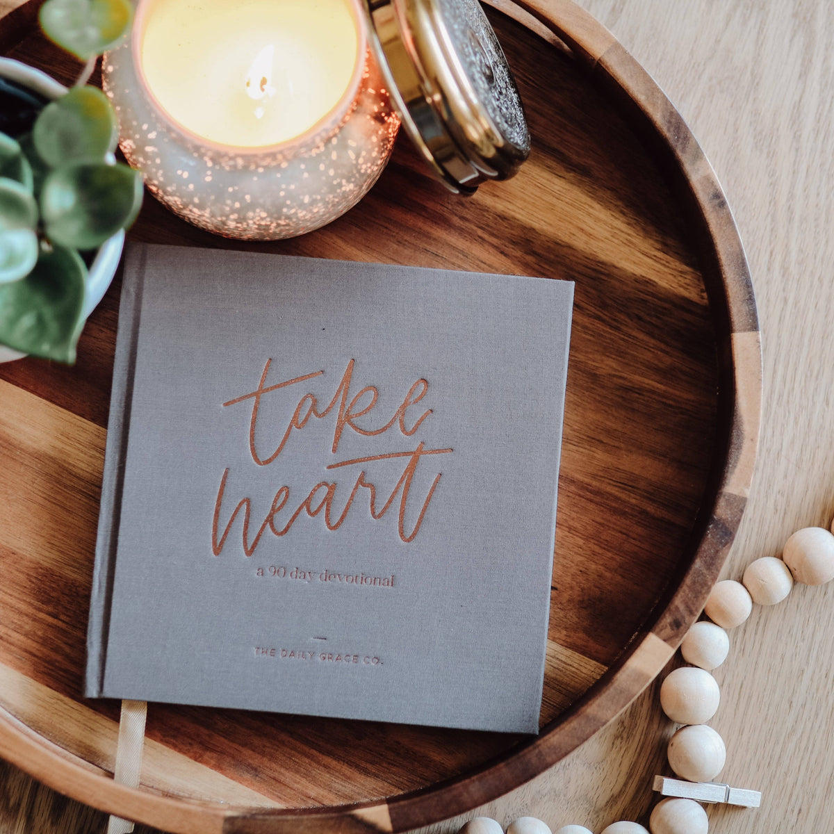 Take Heart devotional book with blue cover and rose gold lettering, styled on wooden tray with candle, succulent, and pearl beads