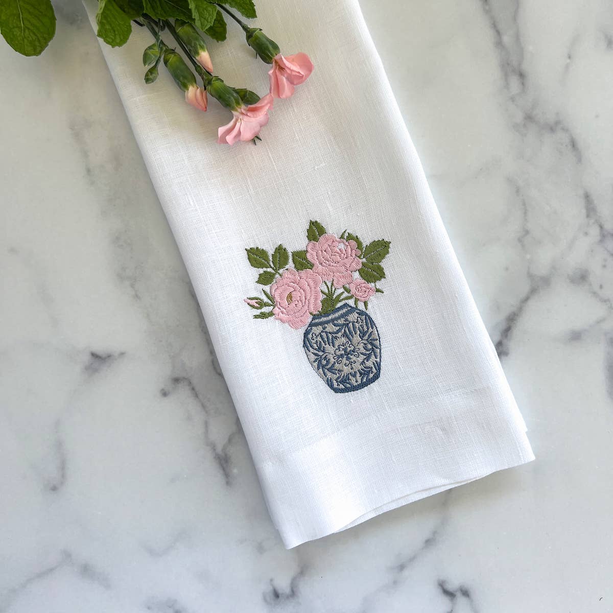 White linen towel with pink rose embroidery in blue vase, fresh pink carnations and greenery arranged on marble background.