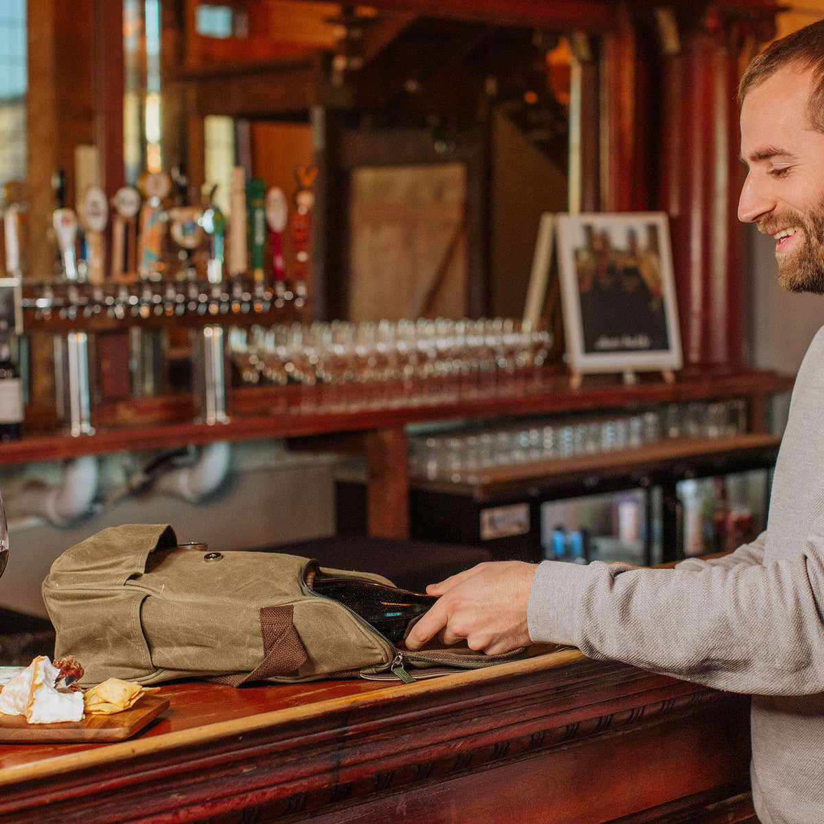 Man opening tan insulated wine tote at wooden bar counter with two glasses of red wine and cheese board