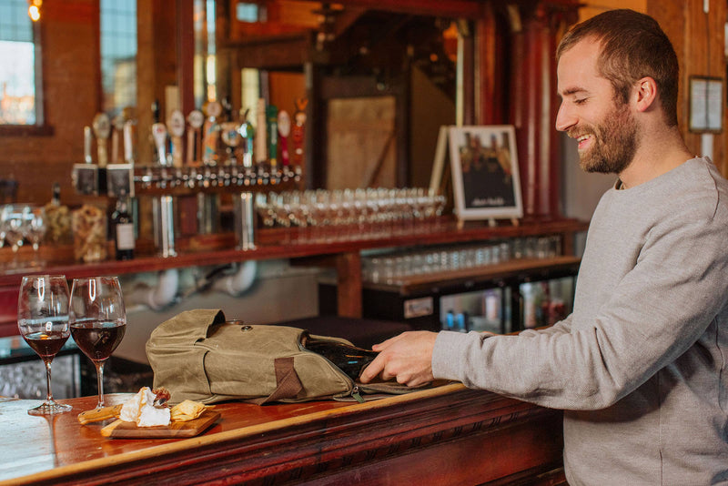 Man opening tan insulated wine tote at wooden bar counter with two glasses of red wine and cheese board