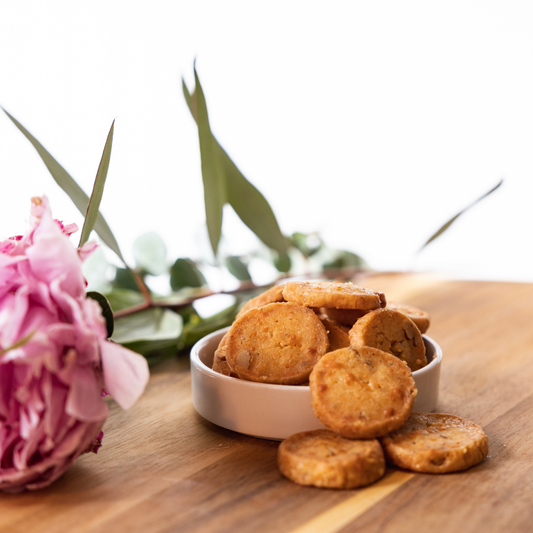 Golden-brown cheddar cookies in a white bowl with scattered crumbles on wood, styled with pink peony and greenery.