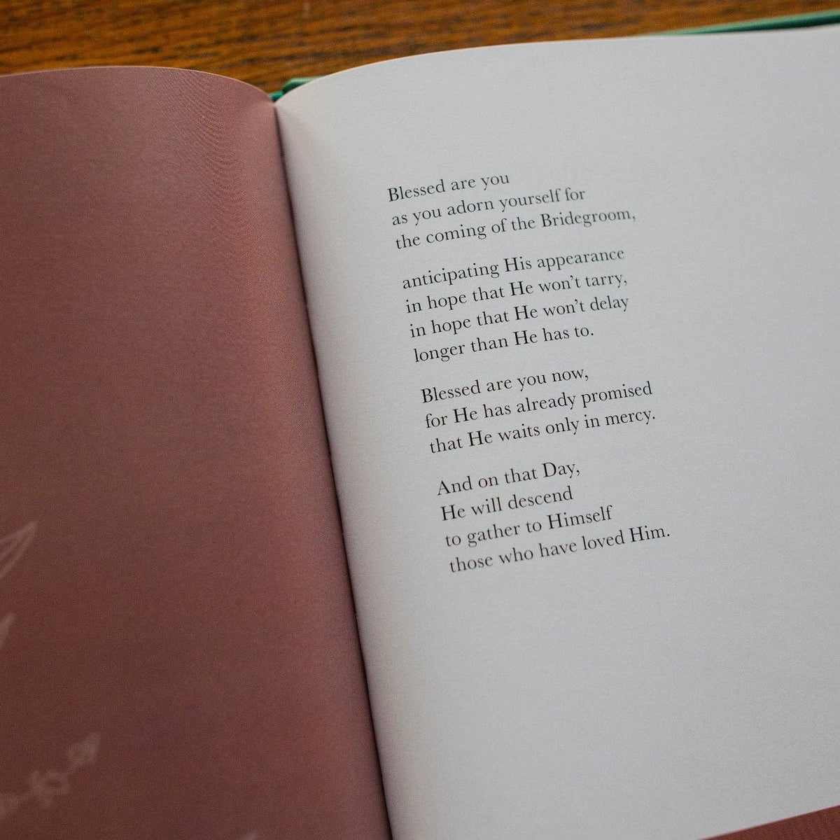 Open prayer book with dusty rose left page and white right page displaying centered text, resting on wooden surface