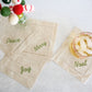 Beige cocktail napkins with Christmas-themed text on a white surface with a glass of iced drink.