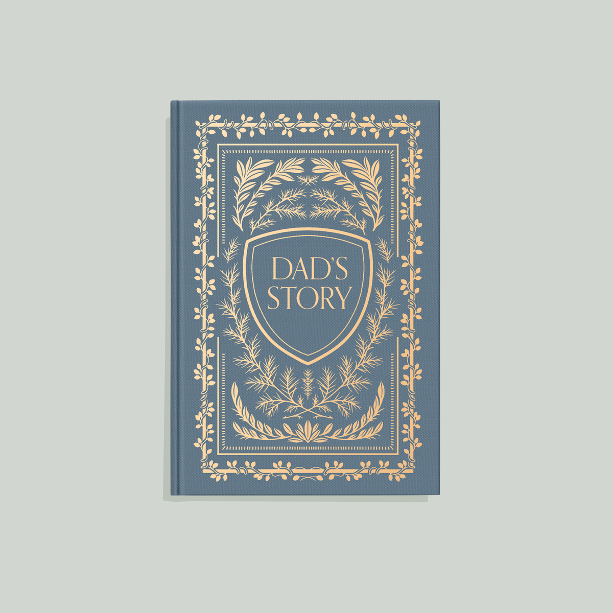 Slate-blue hardcover keepsake journal with ornate gold foil leaf motifs and central "Dad's Story" shield crest