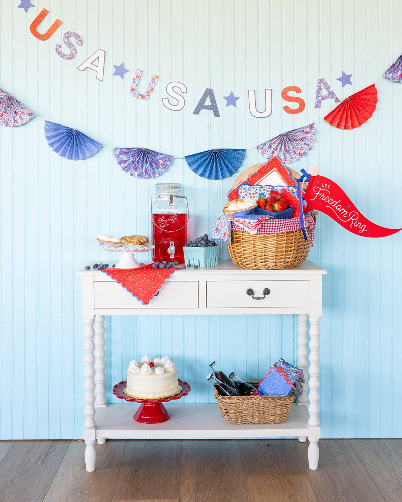Colorful "USA" felt pennant banner with red, white, and purple letters strung across a light blue wall above a white shelf display with patriotic decorations.