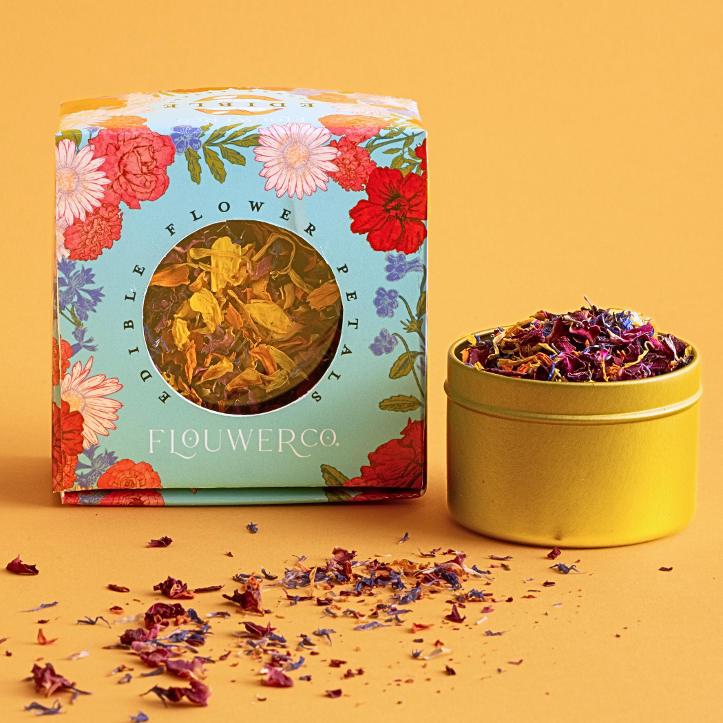 Flouwer Co. edible flower petals in floral-patterned box with circular window, plus gold container and scattered dried petals on yellow background