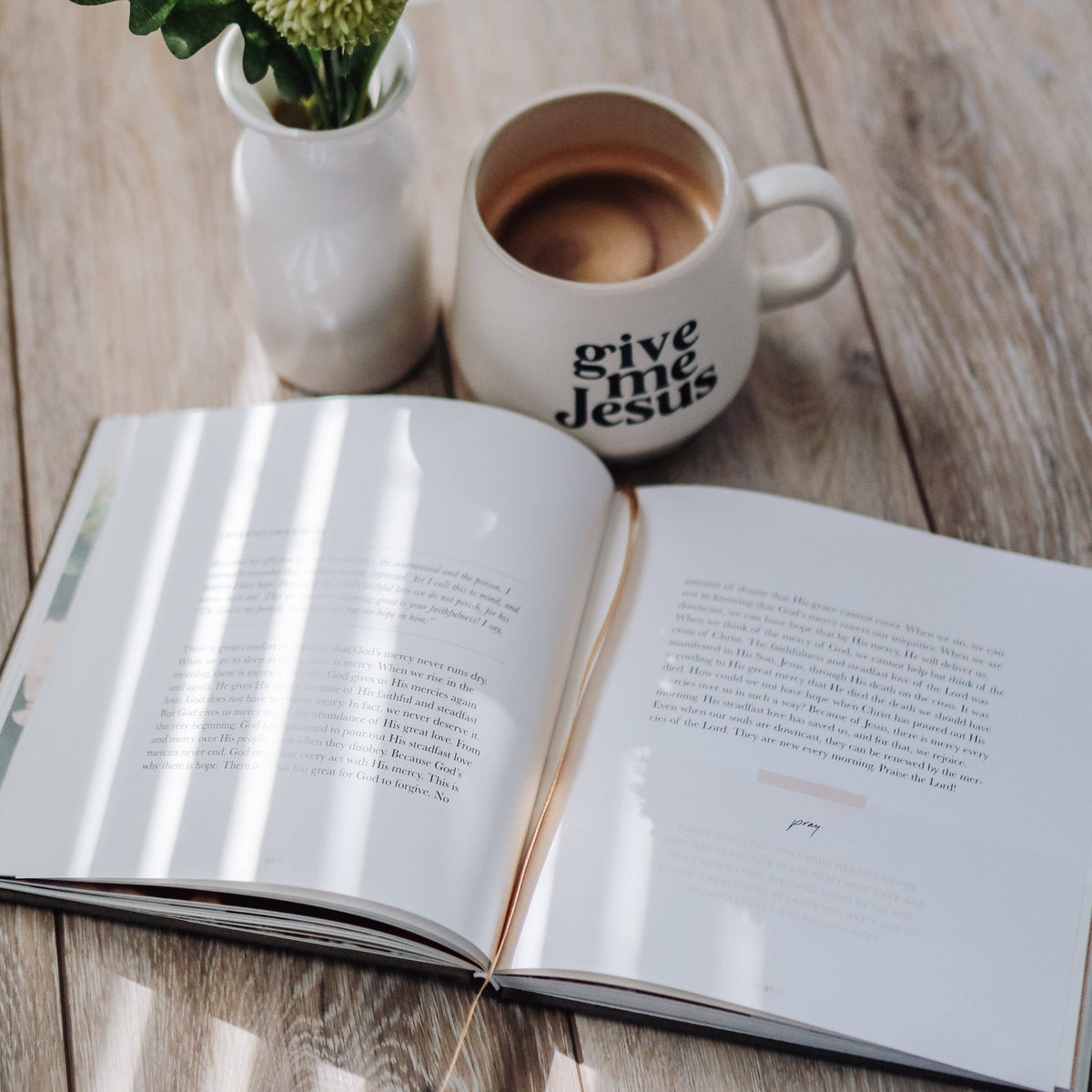 Open devotional book with typed pages beside white mug reading "give me jesus" holding coffee and white vase with green flowers on light wood surface.