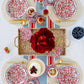 Red floral patterned paper plates arranged on woven placemats with gold flatware, red striped napkins, and fresh rose centerpiece