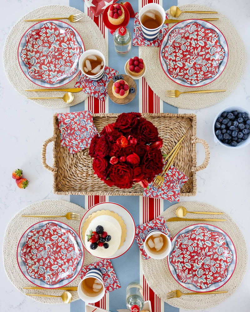 Red floral patterned paper plates arranged on woven placemats with gold flatware, red striped napkins, and fresh rose centerpiece
