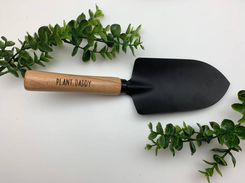 Garden trowel with natural wood handle engraved with "Plant.Daddy" and black metal blade, surrounded by fresh green foliage