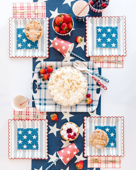 Red, white, and blue star paper cups with checkered napkins and strawberries arranged on patriotic 4th of July party table.