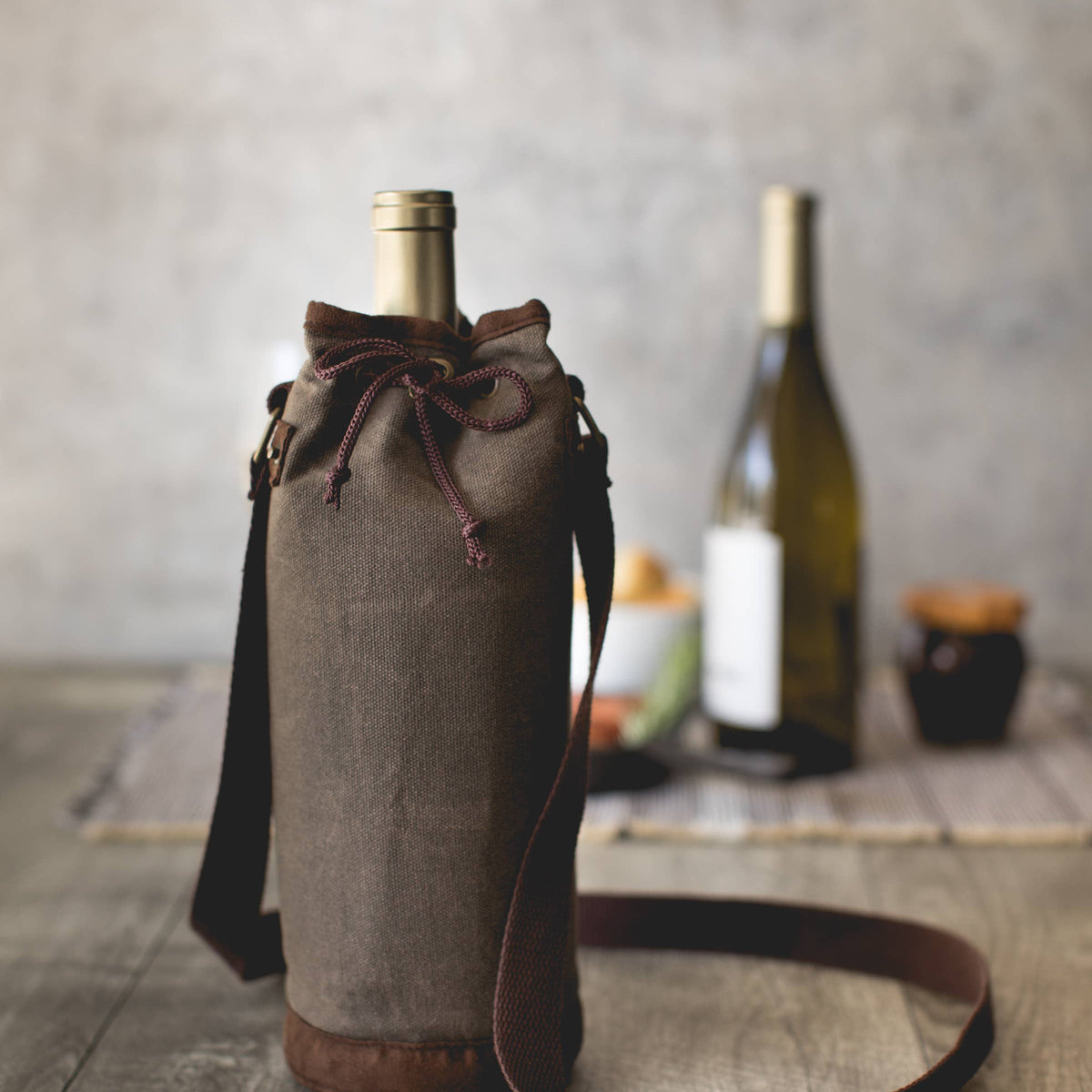Distressed grey waxed canvas wine tote with leather trim, burgundy drawstring closure, and shoulder strap holding a bottle.