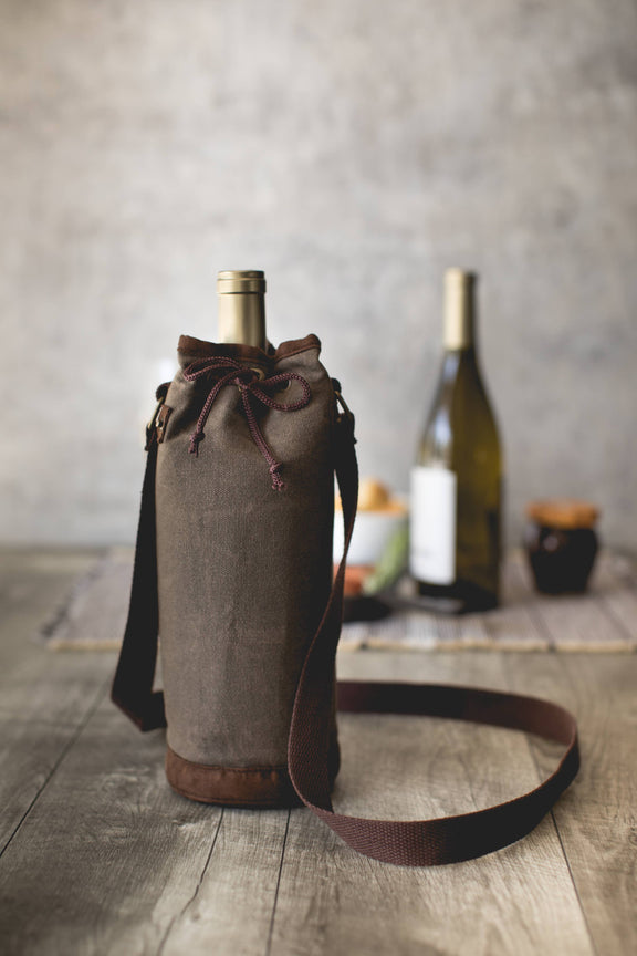 Distressed grey waxed canvas wine tote with leather trim, burgundy drawstring closure, and shoulder strap holding a bottle.