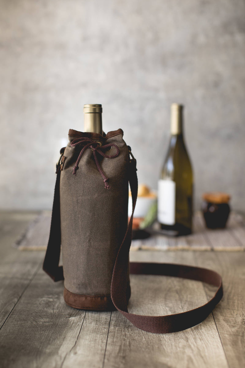 Distressed grey waxed canvas wine tote with leather trim, burgundy drawstring closure, and shoulder strap holding a bottle.