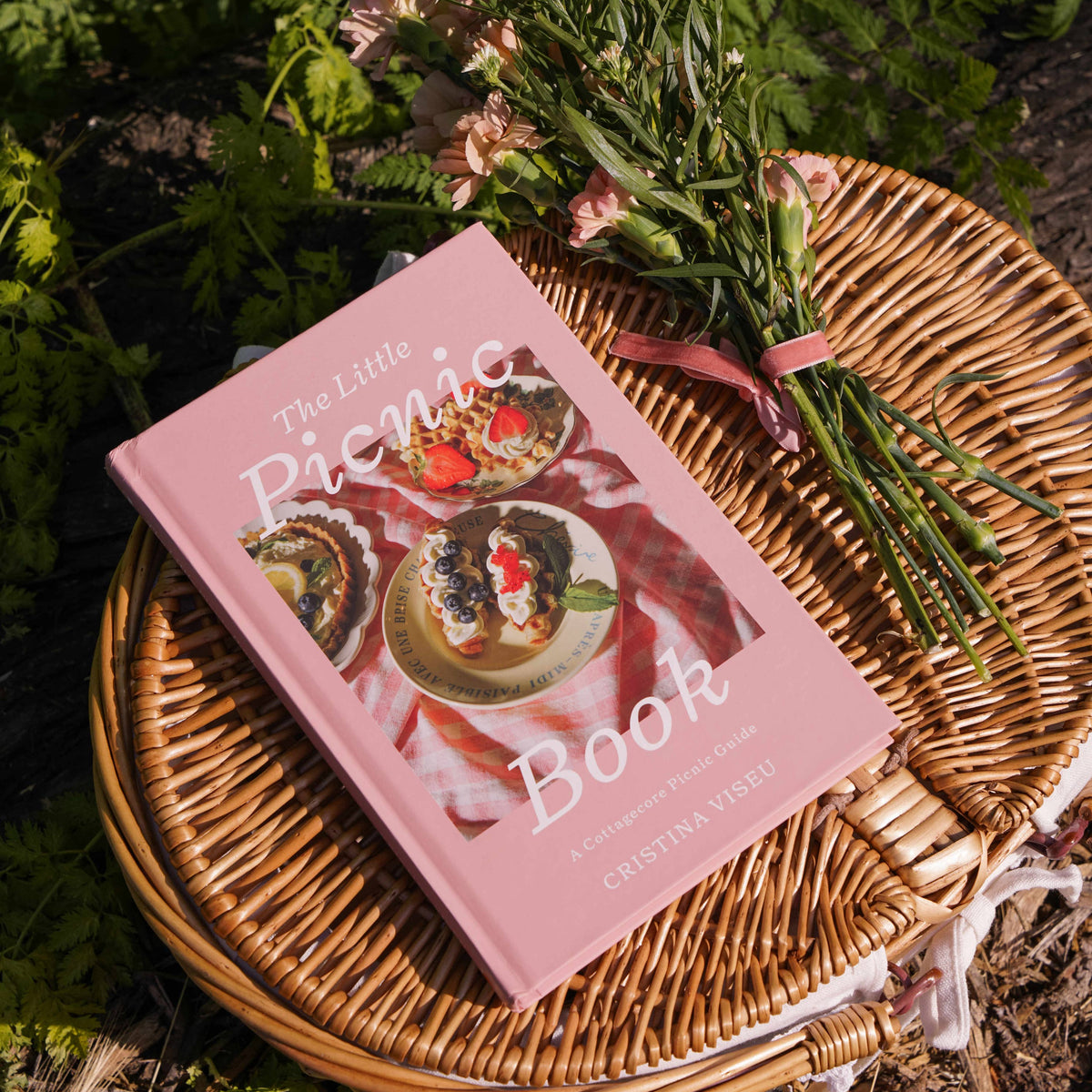 The Little Picnic Book with pink cover on woven wicker plate surrounded by fresh-cut flowers and garden foliage in outdoor picnic setting.