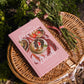 The Little Picnic Book with pink cover on woven wicker plate surrounded by fresh-cut flowers and garden foliage in outdoor picnic setting.