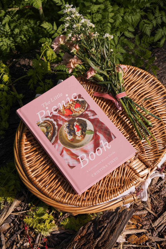 The Little Picnic Book with pink cover on woven wicker plate surrounded by fresh-cut flowers and garden foliage in outdoor picnic setting.