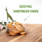 Stack of golden coconut mac shortbread cookies spilling from a white bowl on a wooden surface with fresh green foliage