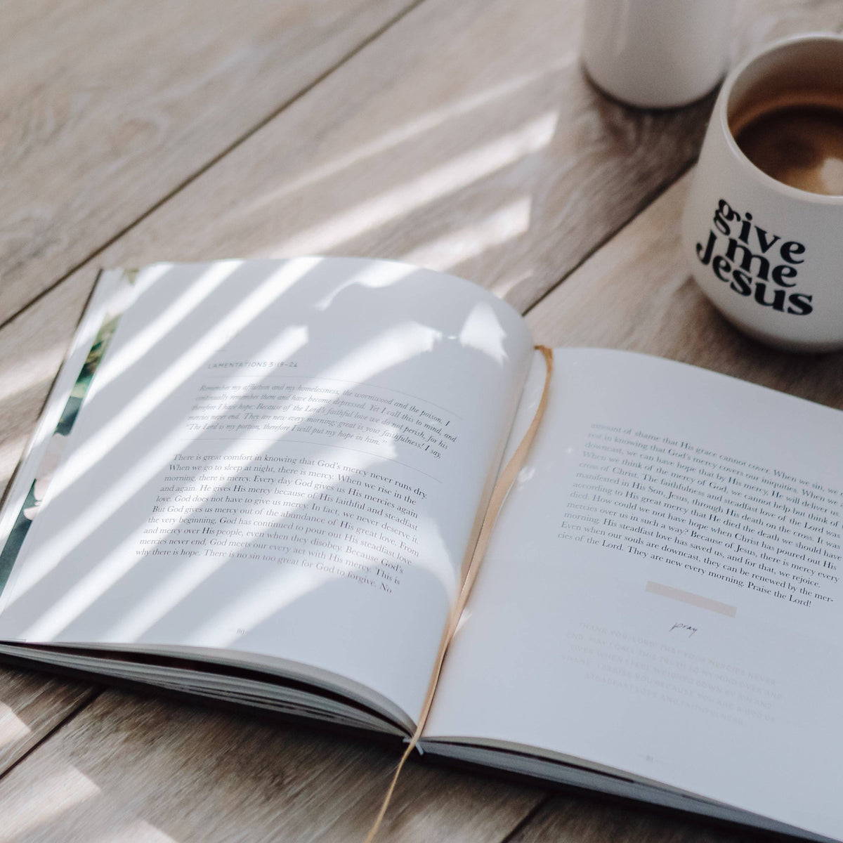 Open devotional book with cream bookmarks on light wood table, white "Give the Jesus" mug and green pen nearby, natural sunlight streaming across