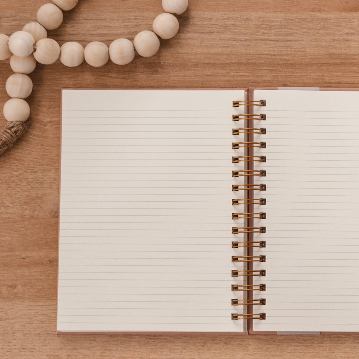 Open spiral-bound journal with cream lined pages, mauve cover, and bronze binding displayed on wood with wooden beads