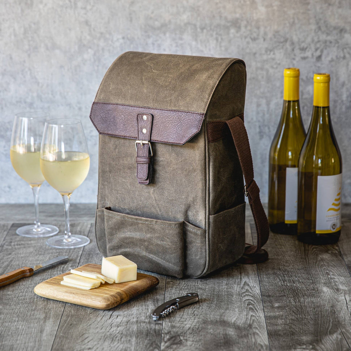 Tan canvas wine tote with burgundy leather flap holding two wine bottles, displayed with glasses of white wine, cheese board, and corkscrew