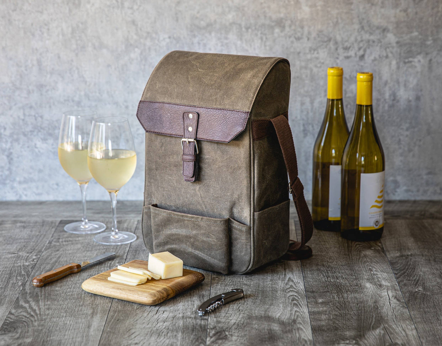 Tan canvas wine tote with burgundy leather flap holding two wine bottles, displayed with glasses of white wine, cheese board, and corkscrew