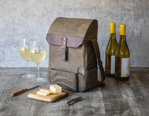 Tan canvas wine tote with burgundy leather flap holding two wine bottles, displayed with glasses of white wine, cheese board, and corkscrew