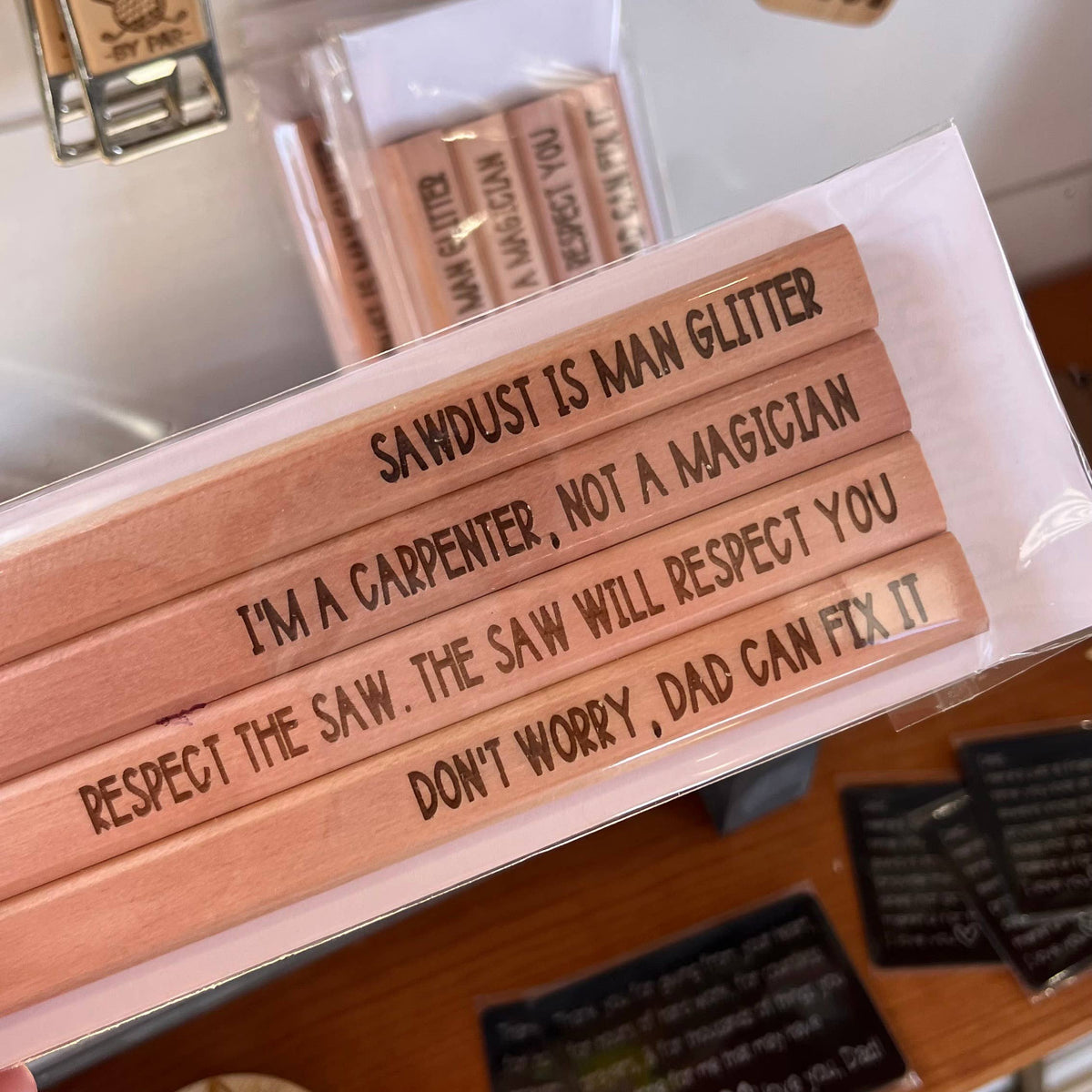Unsharpened flat wooden carpenter pencils with engraved text including "Sawdust is Man Glitter" and "I'm a Carpenter, Not a Magician," displayed with matching wooden keychains.