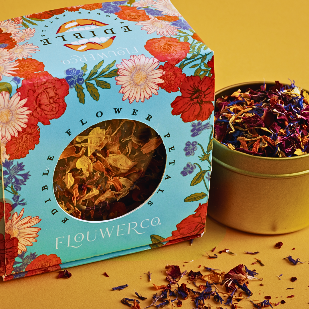 Turquoise box of Flouwer Co. edible flower petals with colorful floral design and gold bowl of dried rose, daisy, and flower petals on mustard background