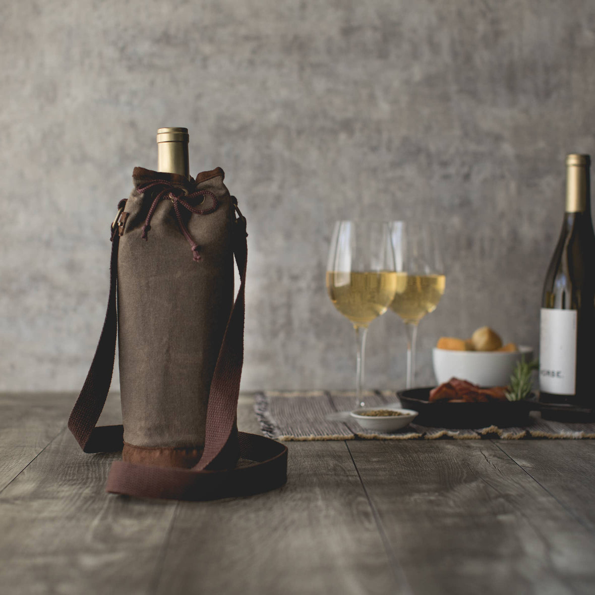 Tan distressed waxed canvas wine tote with burgundy handles and drawstring closure, holding a wine bottle beside wine glasses on wooden surface