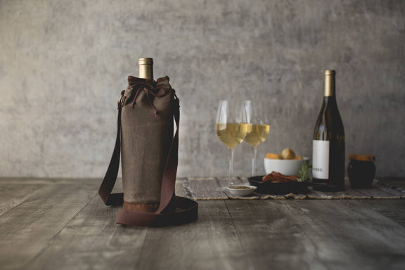Tan distressed waxed canvas wine tote with burgundy handles and drawstring closure, holding a wine bottle beside wine glasses on wooden surface
