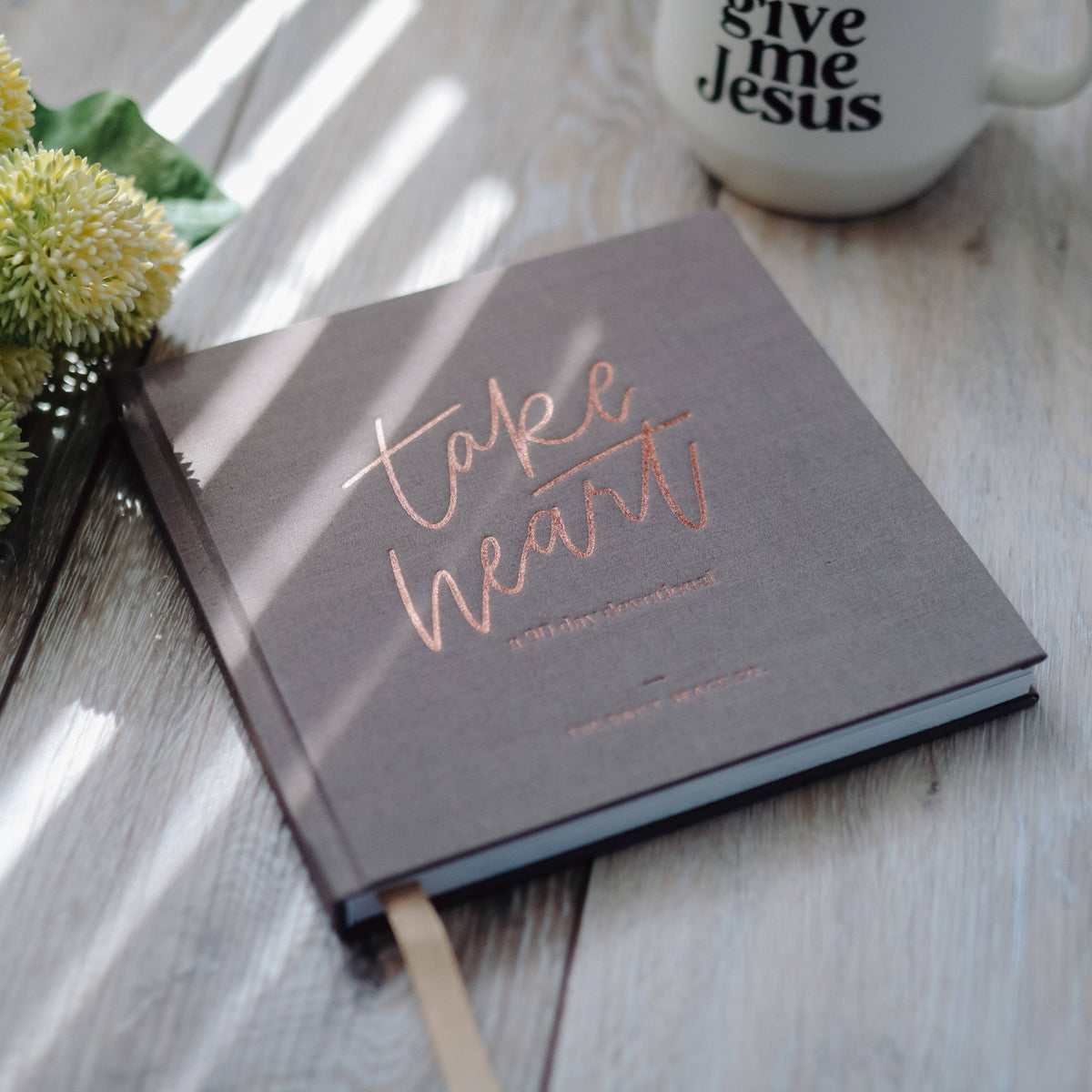 Take Heart 90-day devotional with charcoal cover, rose-gold foil lettering, and cream bookmark on light wood surface beside yellow flower