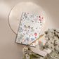 Gardening journal with watercolor wildflower illustrations in coral, blue, and green on cream cover, displayed on round stone plate with gold pen and dried flowers.