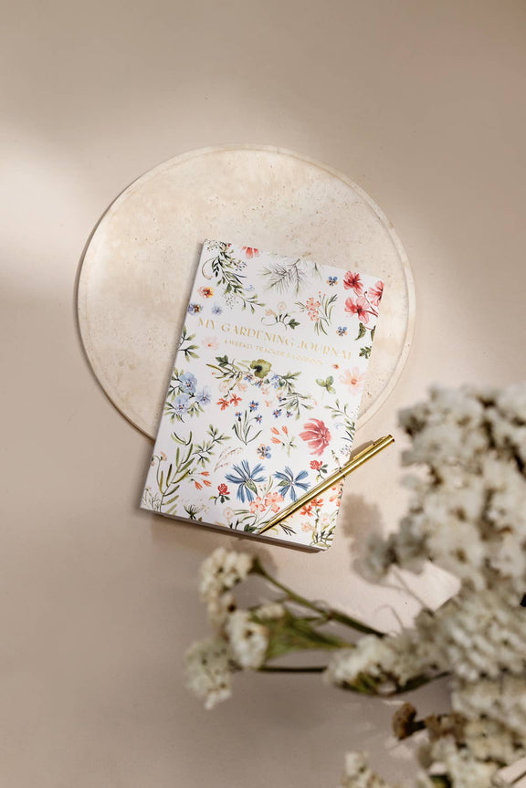Gardening journal with watercolor wildflower illustrations in coral, blue, and green on cream cover, displayed on round stone plate with gold pen and dried flowers.