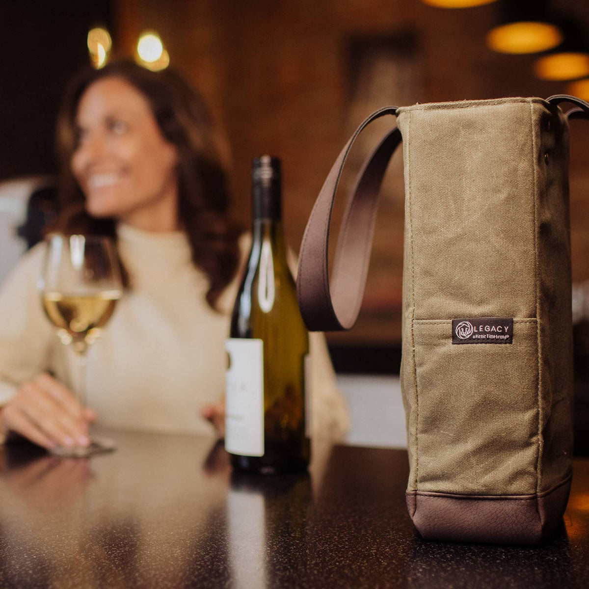 Beige canvas 2-bottle wine tote with brown leather base and handles, displayed on a table at an upscale bar with wine glasses nearby