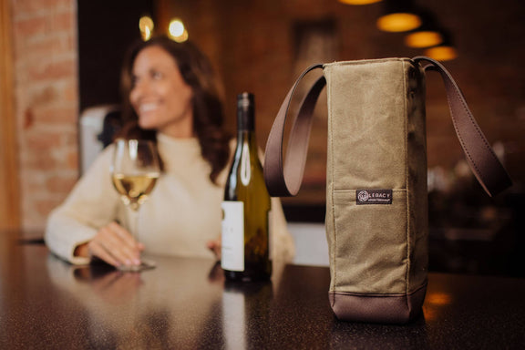 Beige canvas 2-bottle wine tote with brown leather base and handles, displayed on a table at an upscale bar with wine glasses nearby