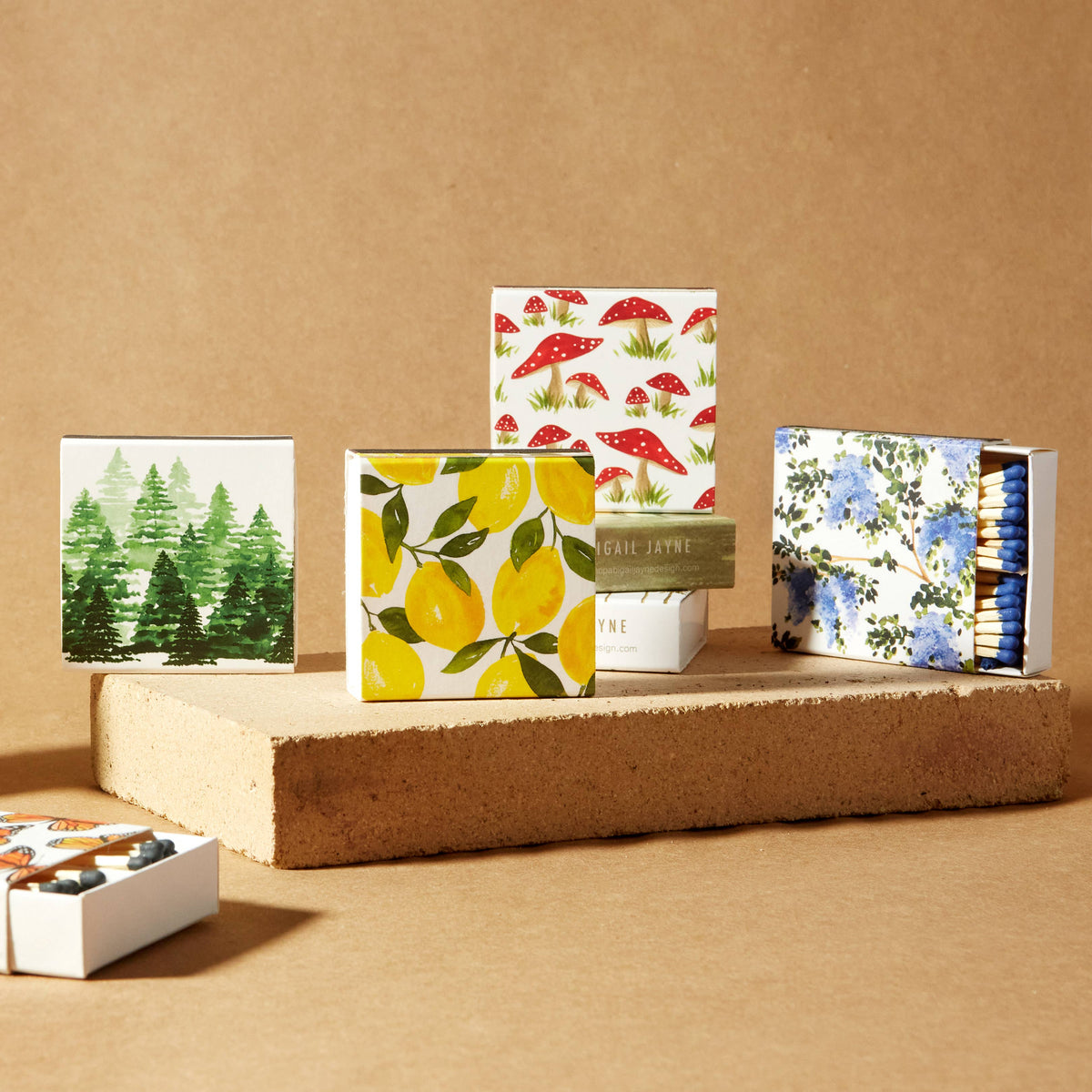 Hand-painted lemon and leaf design on square safety match box with black striker, displayed center stage among coordinating floral and forest-themed match collections on cork platforms
