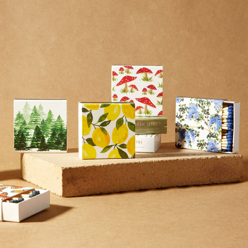 Hand-painted lemon and leaf design on square safety match box with black striker, displayed center stage among coordinating floral and forest-themed match collections on cork platforms