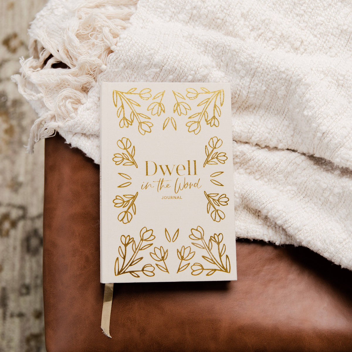 Dwell in the Word Journal with cream cover and gold botanical leaf borders, displayed on brown leather with draped ivory textured fabric.