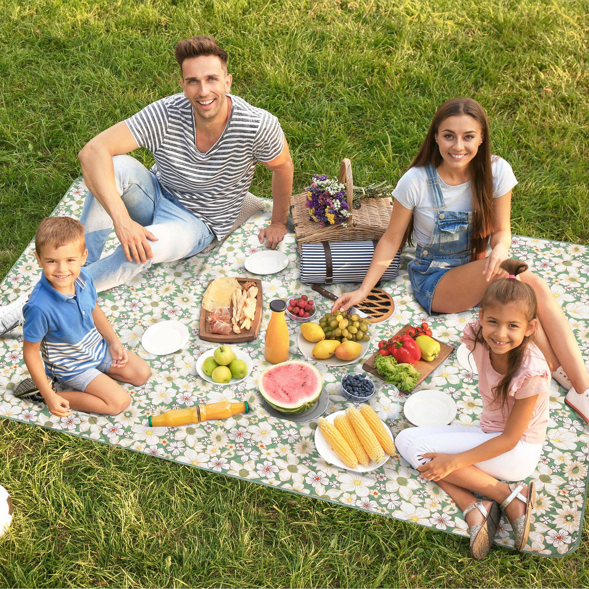 Floral daisy picnic blanket spread on grass with family of four seated around food and drinks for an outdoor meal.