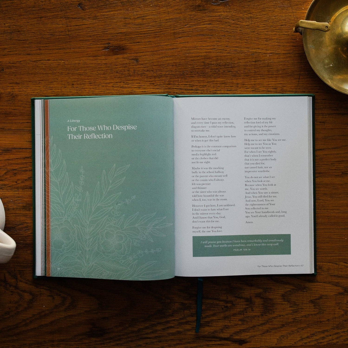 Open prayer book with teal and cream pages displaying liturgical prayers and botanical illustrations, resting on warm wooden surface.