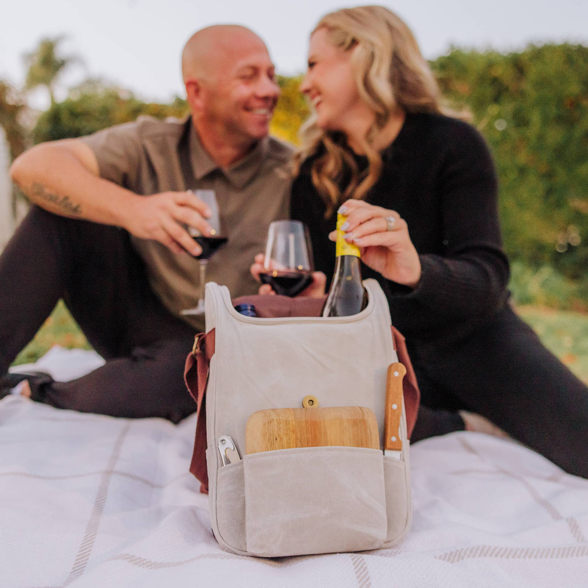 Cream canvas insulated wine cooler with leather handles showing wine bottles and wooden cheese board in front pocket, outdoor picnic setting