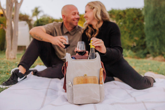 Cream canvas insulated wine cooler with leather handles showing wine bottles and wooden cheese board in front pocket, outdoor picnic setting