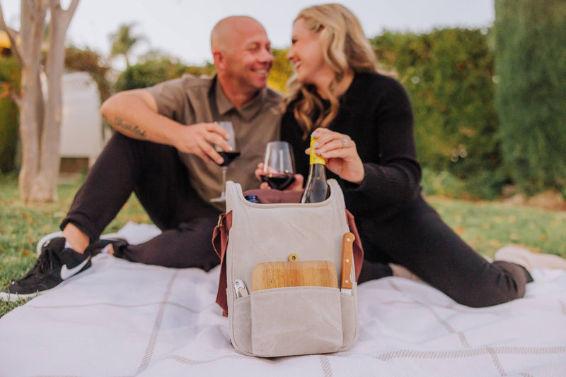 Cream canvas insulated wine cooler with leather handles showing wine bottles and wooden cheese board in front pocket, outdoor picnic setting