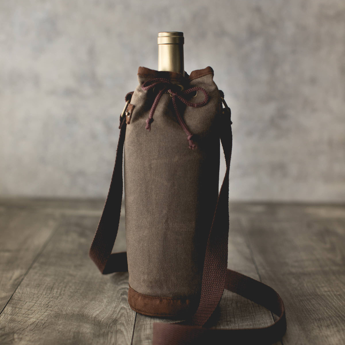 Tan waxed canvas wine bottle carrier with burgundy drawstring closure and adjustable shoulder strap, holding a wine bottle.