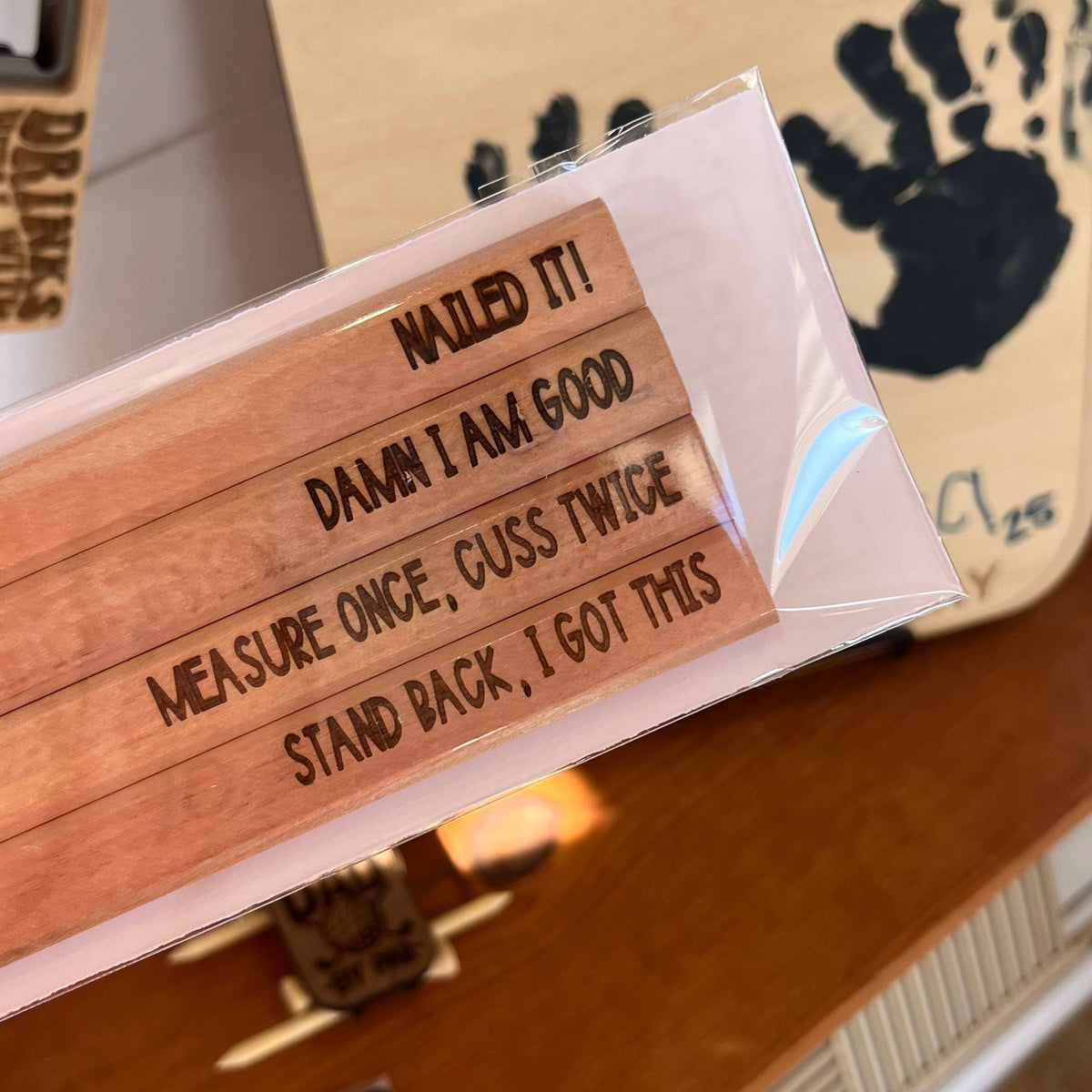 Engraved flat carpenter pencils in natural wood with humorous text phrases, displayed in clear wrapper with "Best Dad" wooden sign background.