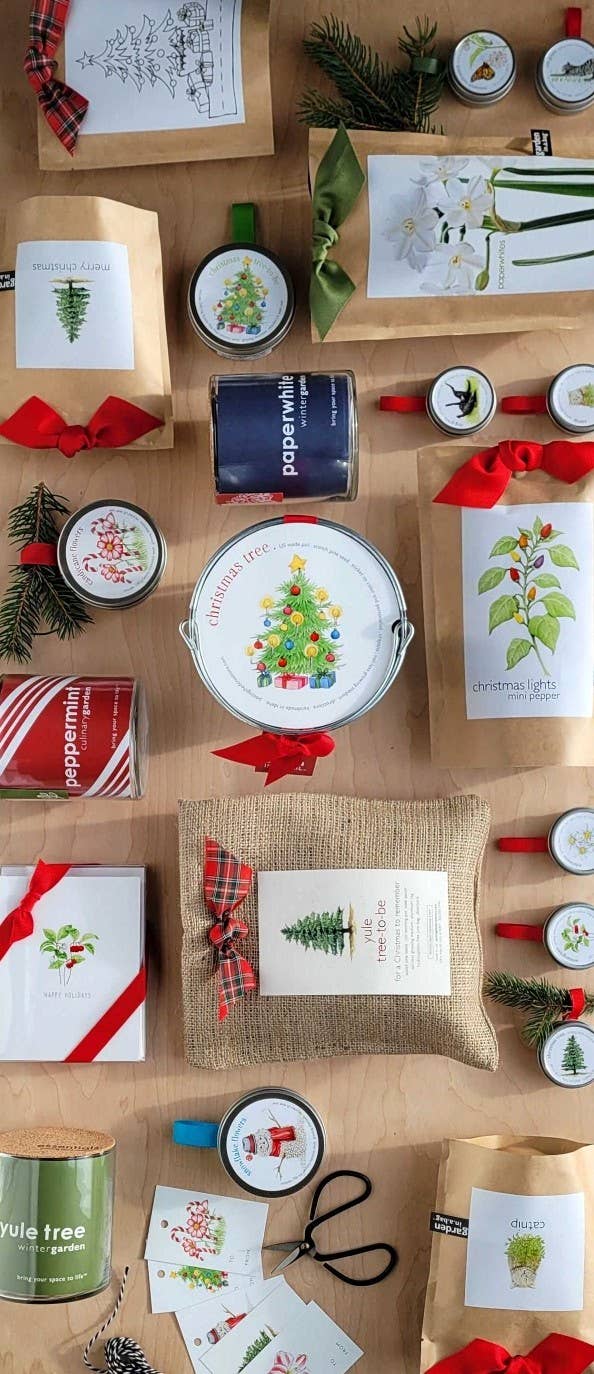 Garden in a Bag paperwhite gift set flat-lay showing seed packets, holiday-printed metal tins with ribbon bows, botanical illustrations, and festive red plaid accents on kraft and burlap