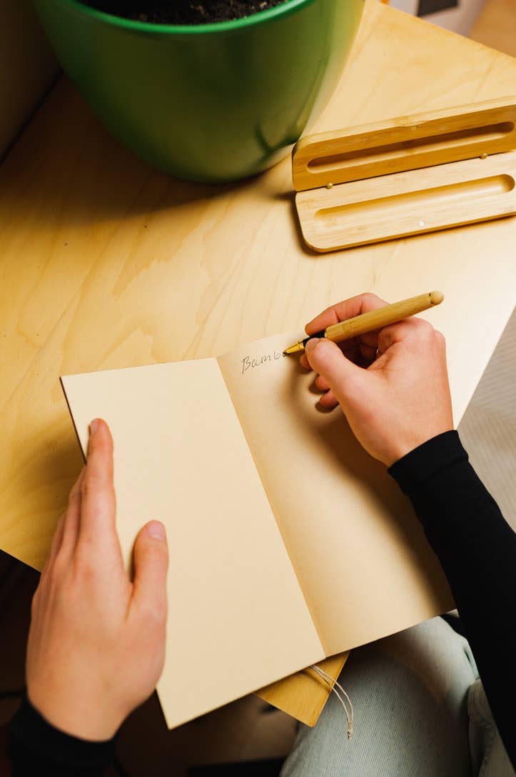 Hand holding bamboo pen writing on white paper with two bamboo pen cases on light wood desk.
